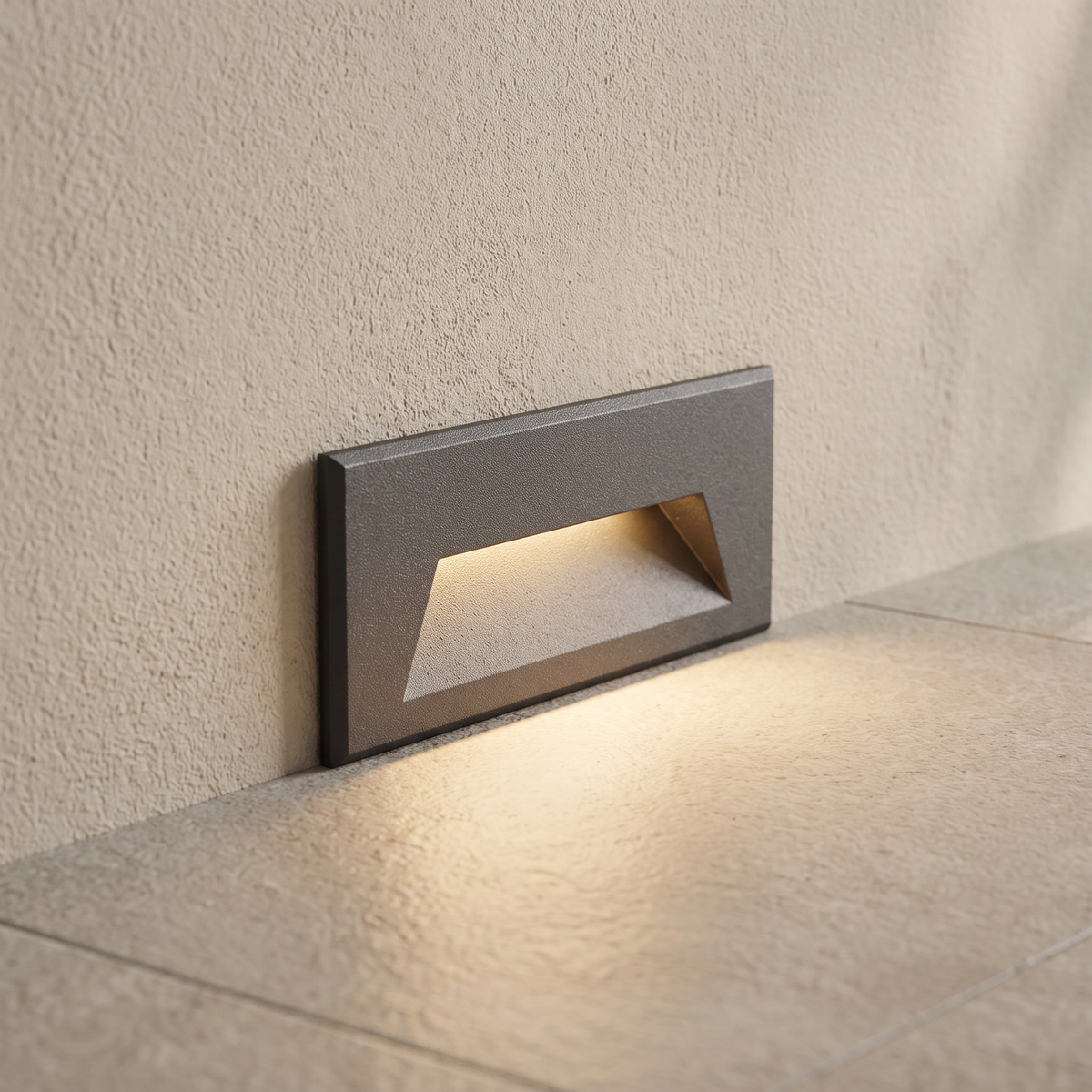 Close-up of one rectangular LED step light recessed low in a light plaster wall, casting a warm wash of light onto the floor