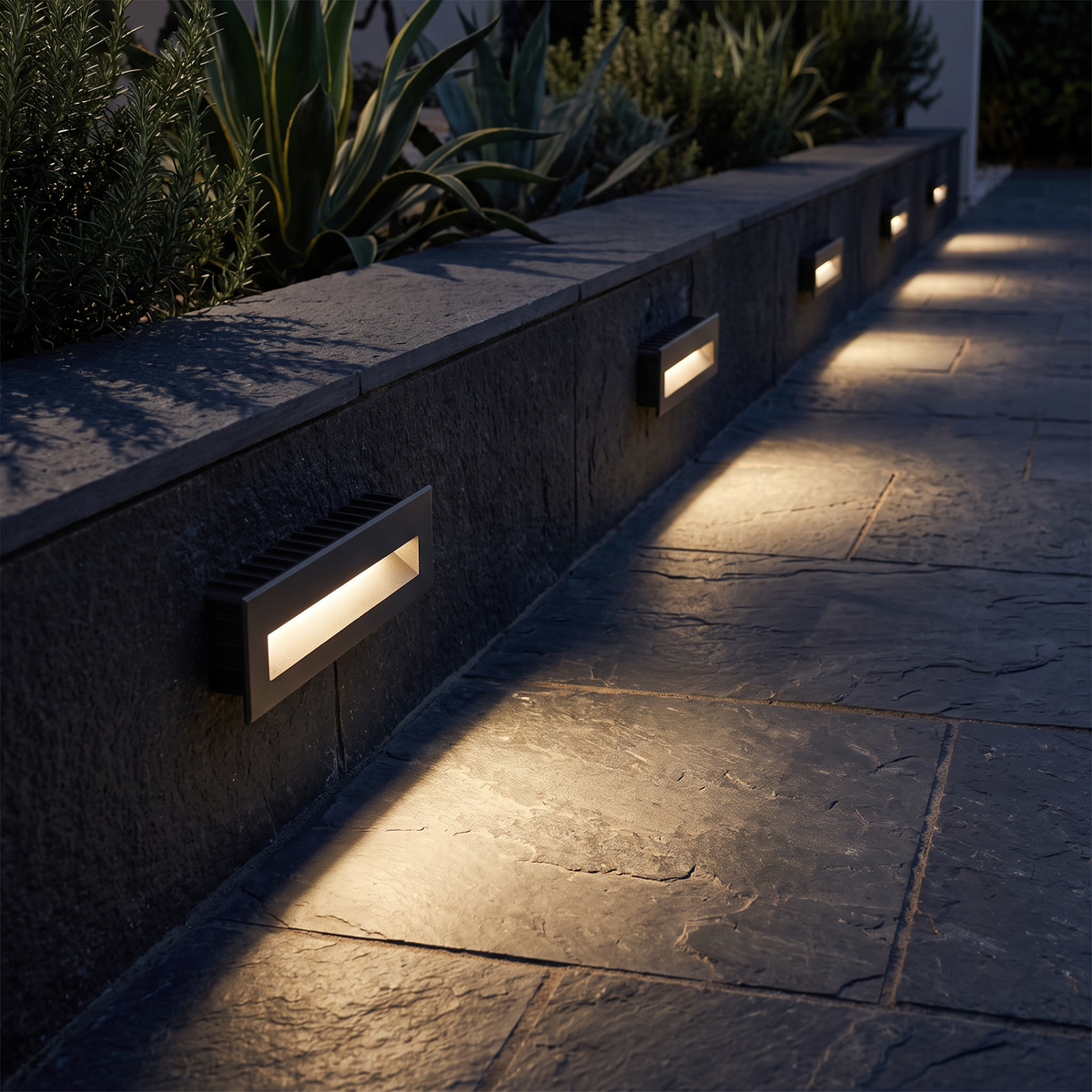 Long rectangular LED step lights recessed along the wall of a villa entrance pathway at night, lighting the paving