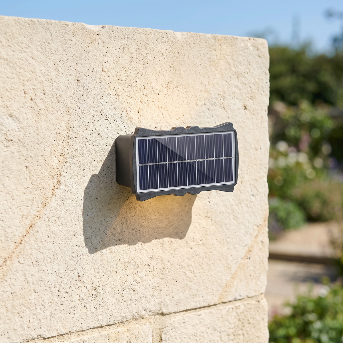 The solar up and down wall light mounted on a light stone garden wall on a sunny day, solar panel on top visible